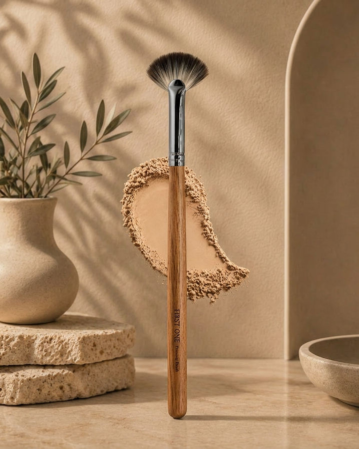 Makeup brush with a wooden handle and silver ferrule, held above a small round mirror with beige powder on a neutral background.  Elegant flatlay featuring The Golden Doe fan highlighter brush alongside highlighter powder. Text overlay: ‘Precision Highlight & Glow Brush — seamless even coverage’. Clean aesthetic, warm tones, professional beauty tool.