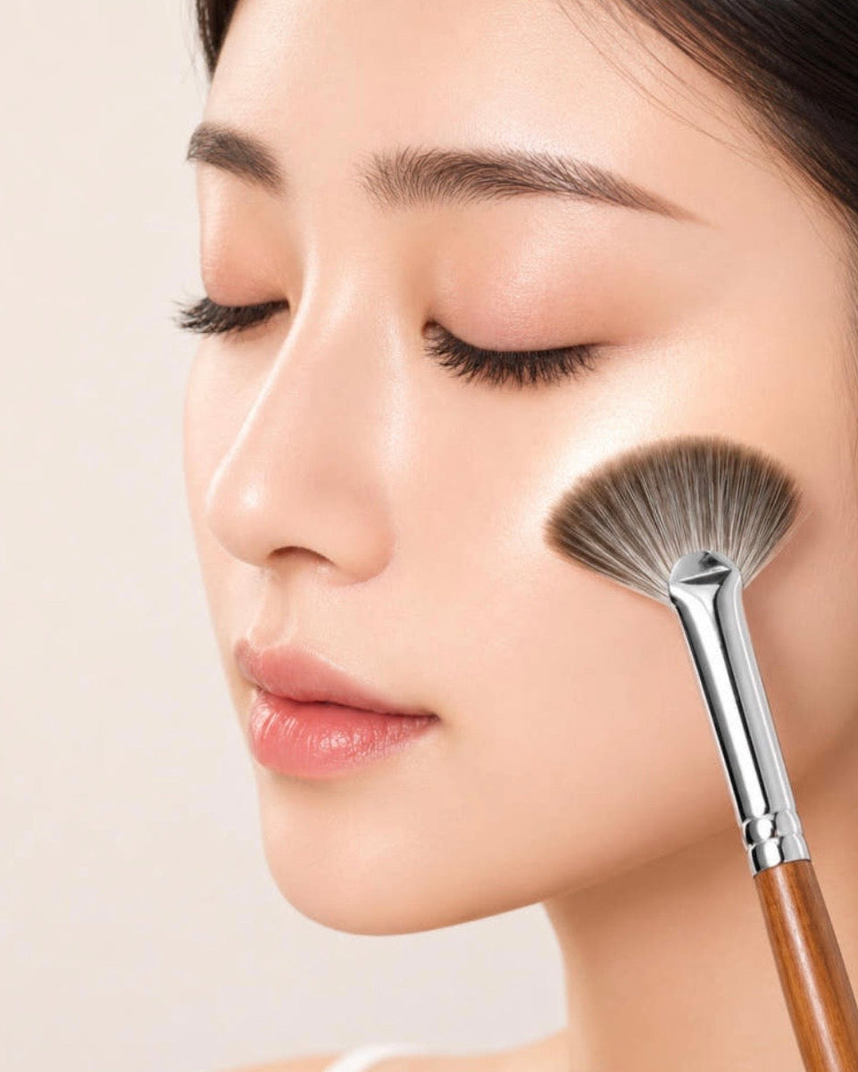 Close-up of a woman's face with closed eyes as a makeup brush applies cream product to her cheek, set against a soft beige background.

Soft fan brush sweeping across cheekbones to apply champagne highlighter. Gentle glide creating seamless natural glow, no streaks or fallout. Hypoallergenic ultra-soft bristles, smooth skin-like finish.