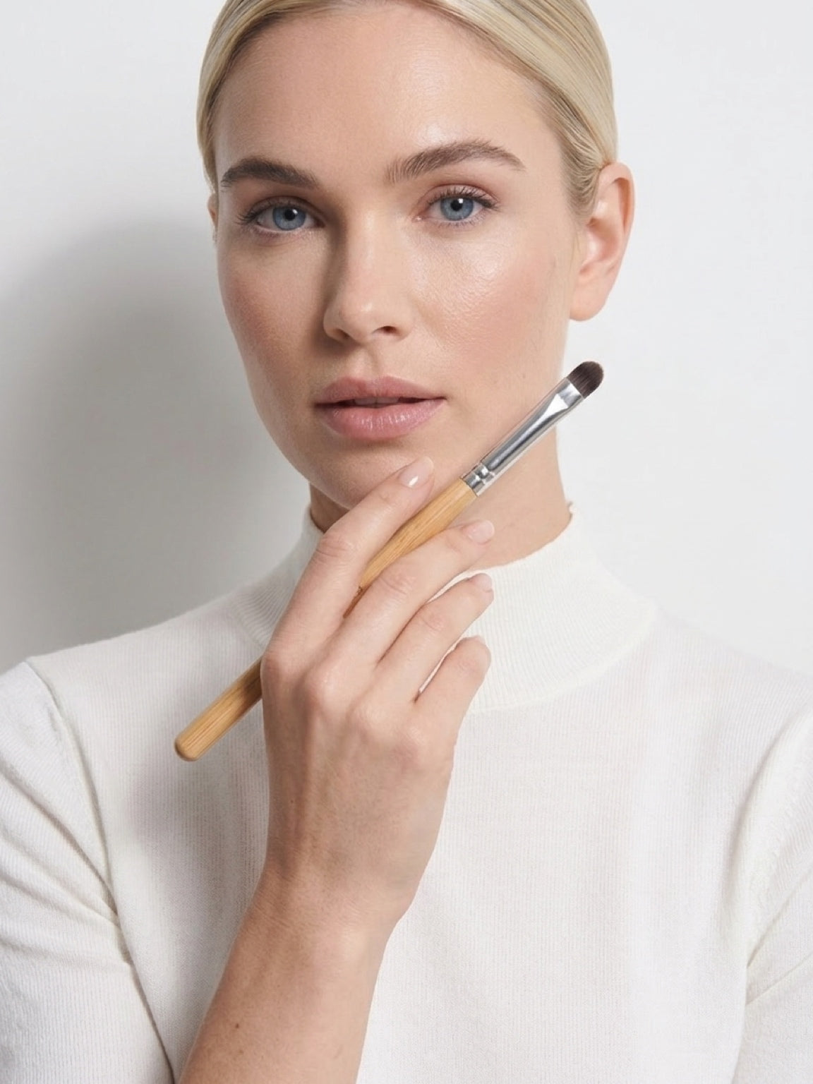 A close-up of a woman holding a makeup brush near her face, wearing a cream mock-neck top against a light gray background.  The Lip Sculptor Brush — slim tapered tip, precision lip brush. Text: ‘Sculpts, defines, creates clean edges effortlessly’. Soft yet firm bristles, professional design. £12.00.