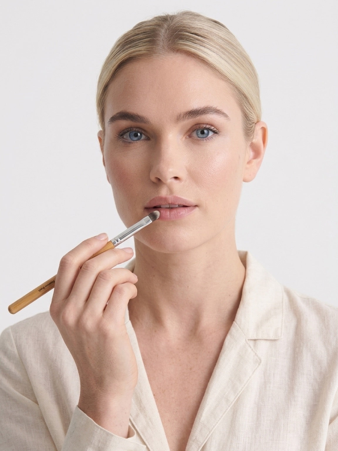 A blonde woman with blue eyes wearing a light beige linen shirt, holding a makeup brush to apply product to her lips, against a plain light gray background. Slim tapered brush shaping lip corners and edges with precision. Smooth colour application, crisp defined outline. Blends beautifully — sheer to full coverage. Works with gloss, cream, matte, and balm.
