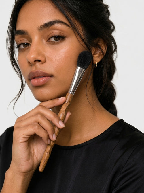 A close-up of a woman holding a makeup brush against her cheek, wearing a black top and small gold hoop earrings, photographed against a light gray background. Small precision blush brush sweeping across cheek, kissing colour into skin. Soft natural flush, seamless skin-like finish. Buildable sheer to full coverage. Black fox hair bristles — impossibly soft, luxurious feel.