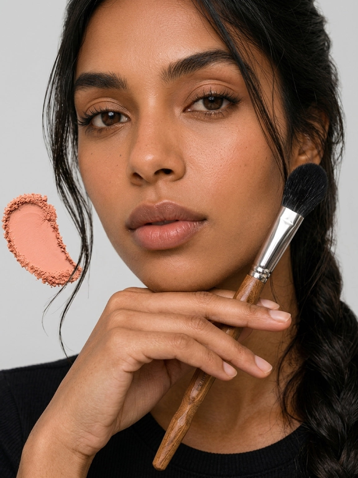 A close-up of a woman holding a makeup brush near her face, with a peach-colored makeup swatch graphic on the left side, set against a plain light gray background. Close-up of brush head showing dense, ultra-soft black fox hair bristles. Text: ‘Exceptionally soft — seriously pigmented. 9mm precise head’. Fine quality fibres, compact shape, luxurious craftsmanship.