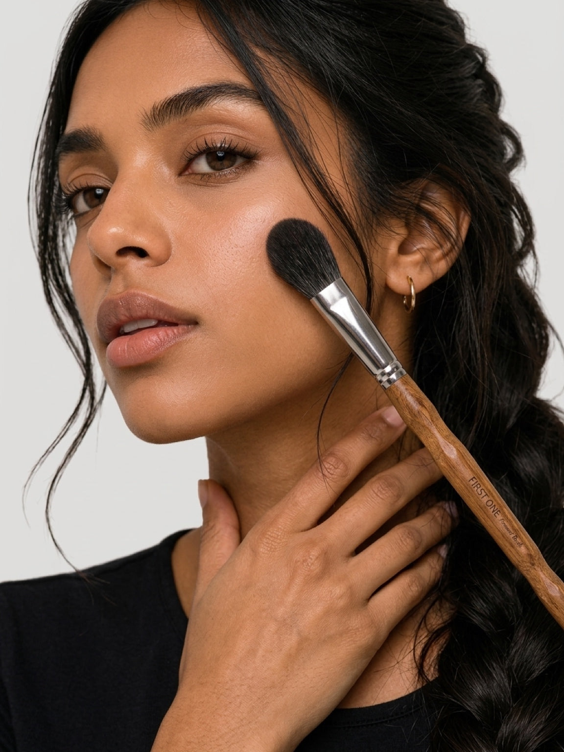 A close-up lifestyle shot of a woman with dark hair applying makeup with a makeup brush against a light gray background. lush applied with Black Fox Brush — soft rose flush, natural ‘I woke up like this’ glow. Text: ‘Your secret weapon for effortless cheek colour’. Powder blush blending beautifully, no harsh edges, warm luminous finish.