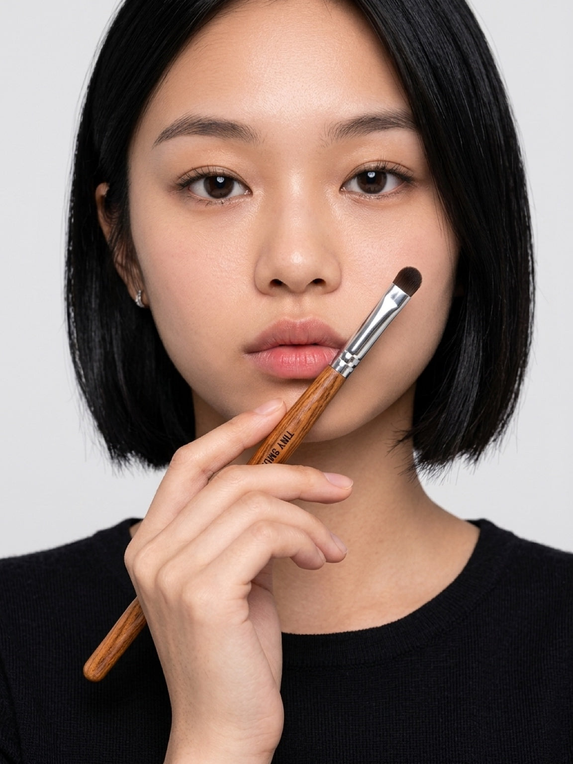 A close-up of a woman with a short black bob holding a makeup brush near her face, wearing a black top against a light gray background. Flat shadow brush pressing colour smoothly across eyelid. Rich saturated pigment deposited evenly, no fallout. Precise controlled application — stays on lid, out of crease. Soft seamless wash of colour, professional finish.