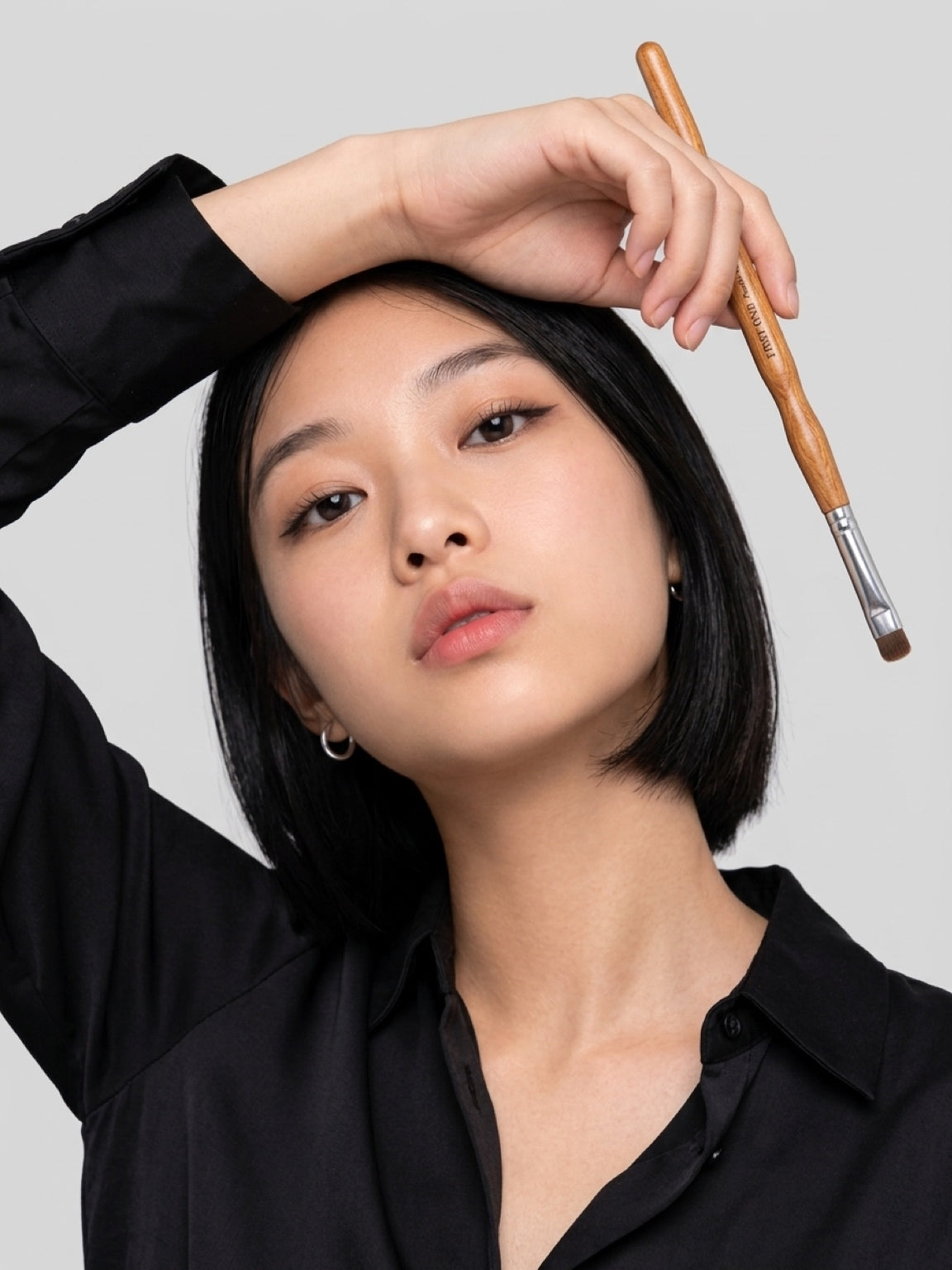 A close-up portrait of a woman with short black hair wearing a black collared shirt, holding a makeup brush near her head against a light gray background. Close-up of brush head showing flat, densely packed premium pony hair bristles. Text: ‘Premium pony hair — grips pigment, no fallout’. Compact 7.5mm size, fine natural fibres, expert craftsmanship.