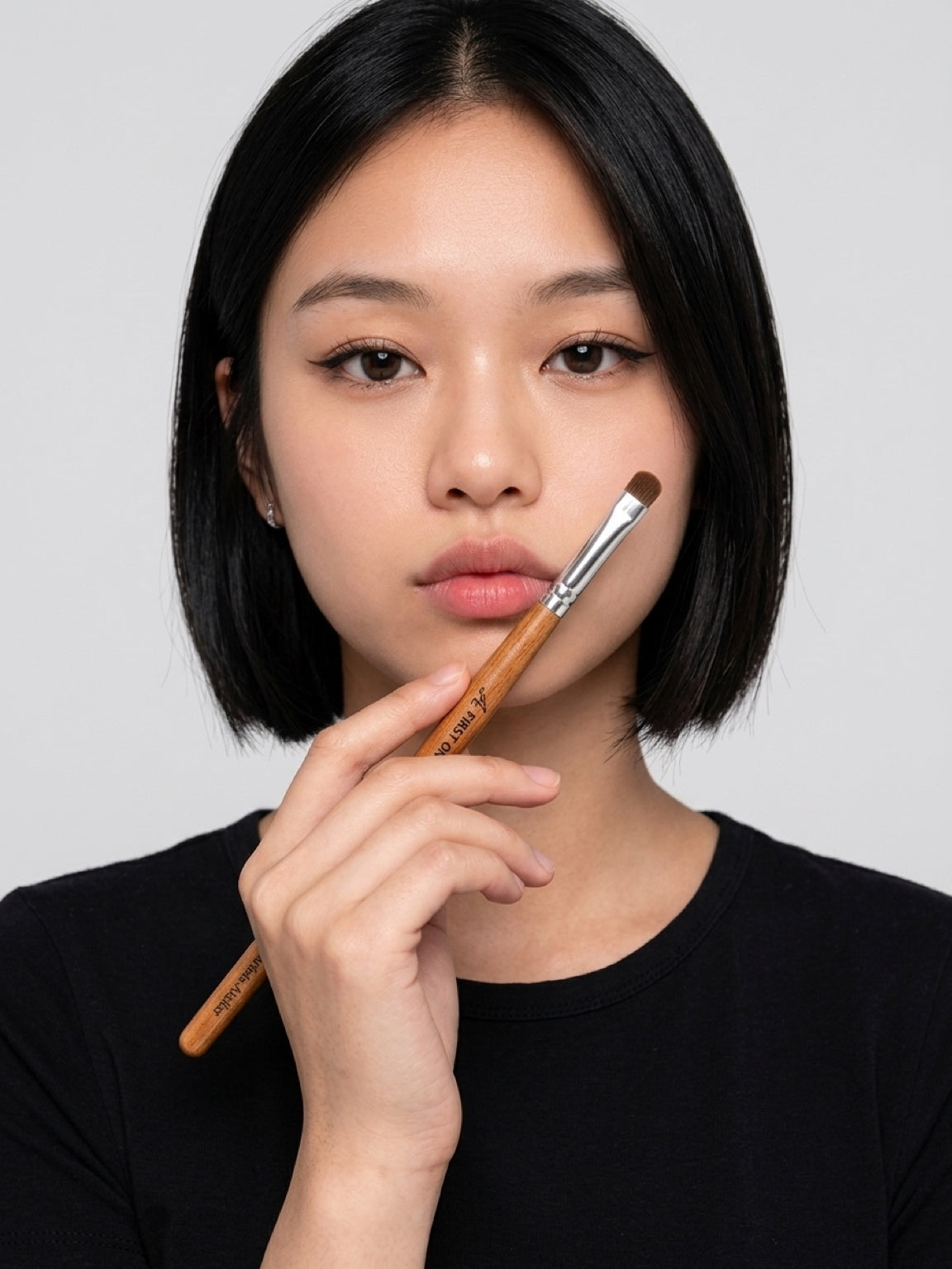 A close-up of a woman with a short black bob holding a makeup brush against her cheek, wearing a black top against a light gray background. Eyeshadow swatch demonstrating rich saturated colour payoff — matte, shimmer, and glitter shades. Text: ‘From sheer wash to intense colour — all formulas work beautifully’. Even pigment deposit, no fading, vibrant true-to-pan colour. Shadow Moth Eyeshadow Brush, Detail & Blending Eye Brush, Artist Atelier, premium pony hair bristles, flat dense head, 7.5mm precision, 