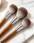 Three makeup brushes with light brown wooden handles and silver ferrules, arranged diagonally on a white marble surface with a small beige dish in the background.