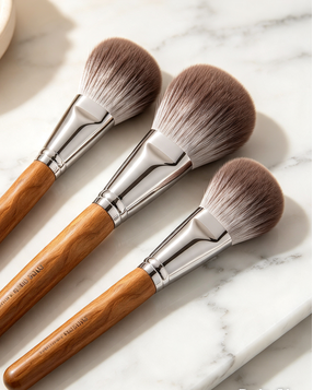 Three makeup brushes with light brown wooden handles and silver ferrules, arranged diagonally on a white marble surface with a small beige dish in the background.